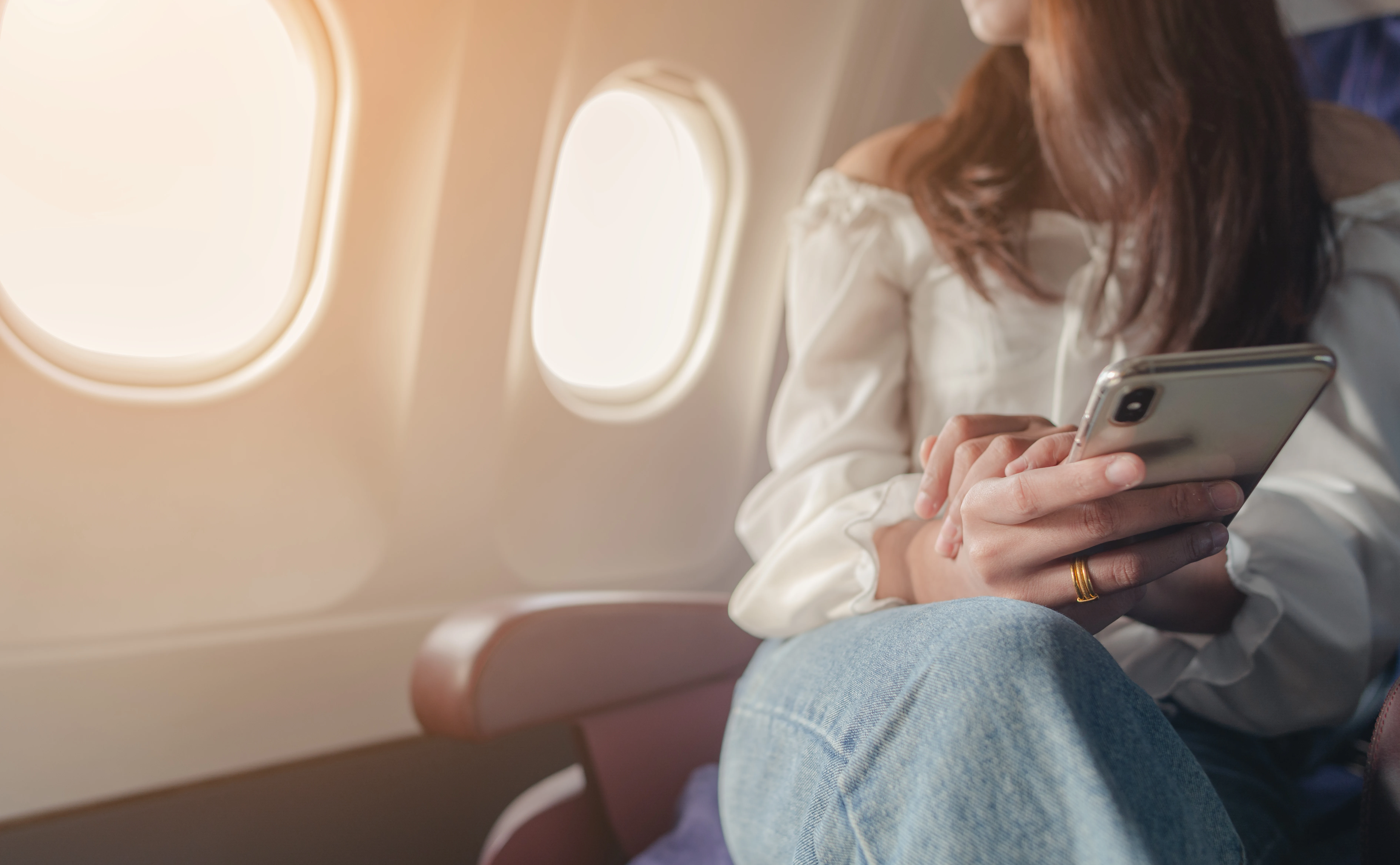 A young woman sits looking out the window of a plane, phone in hand.